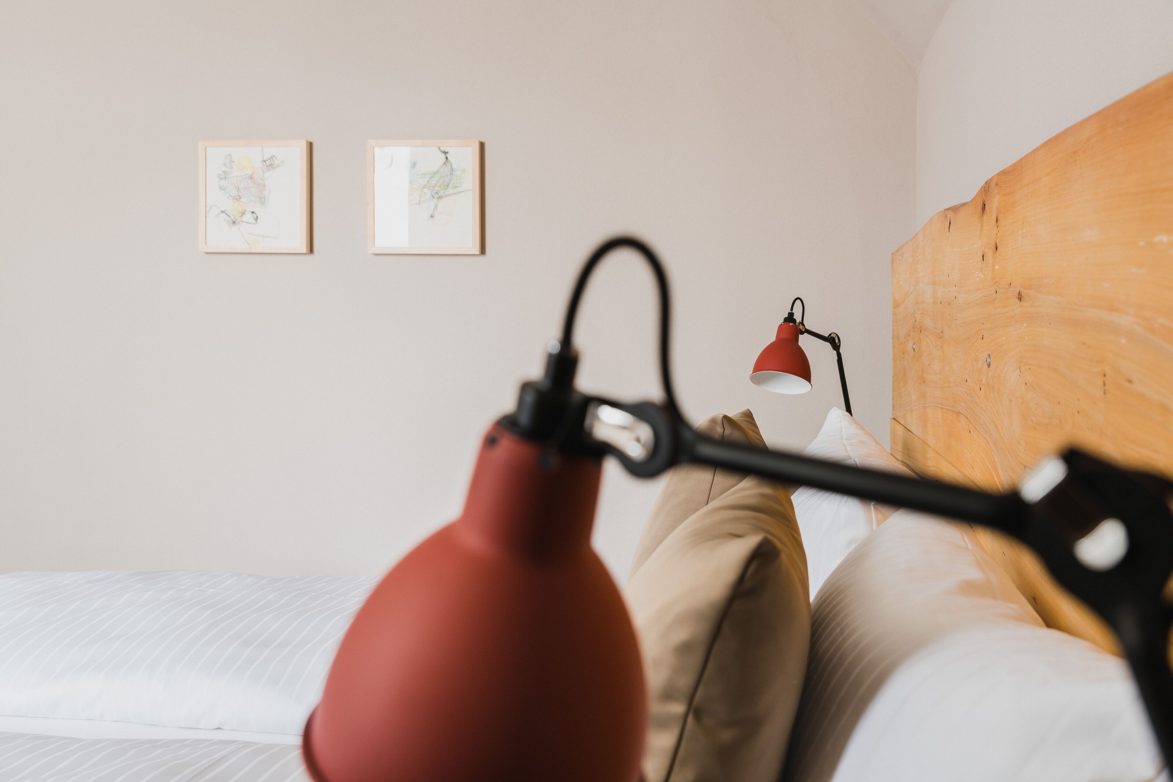 Cama com roupa de cama branca, abajur vermelho e duas obras de arte emolduradas na parede.