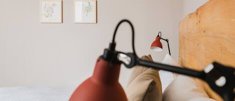 Una cama con ropa de cama blanca, una lámpara roja y dos cuadros en la pared.