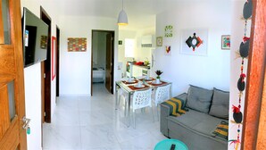 A modern living room with a sofa, dining table, and chairs. There is a kitchen area in the background with a refrigerator and a sink. - RENT apartments in Porto de Galinhas, fully equipped and new! (Porto de Galinhas)