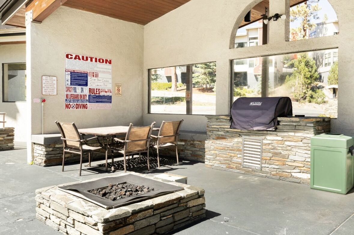 Un patio exterior con un hogar de piedra, una mesa con sillas, una parrilla de barbacoa y vistas a través de amplias ventanas.