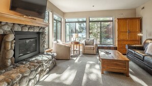 A living room with a stone fireplace, a television mounted above it, a leather sofa, a wooden coffee table, and a large wooden cabinet.