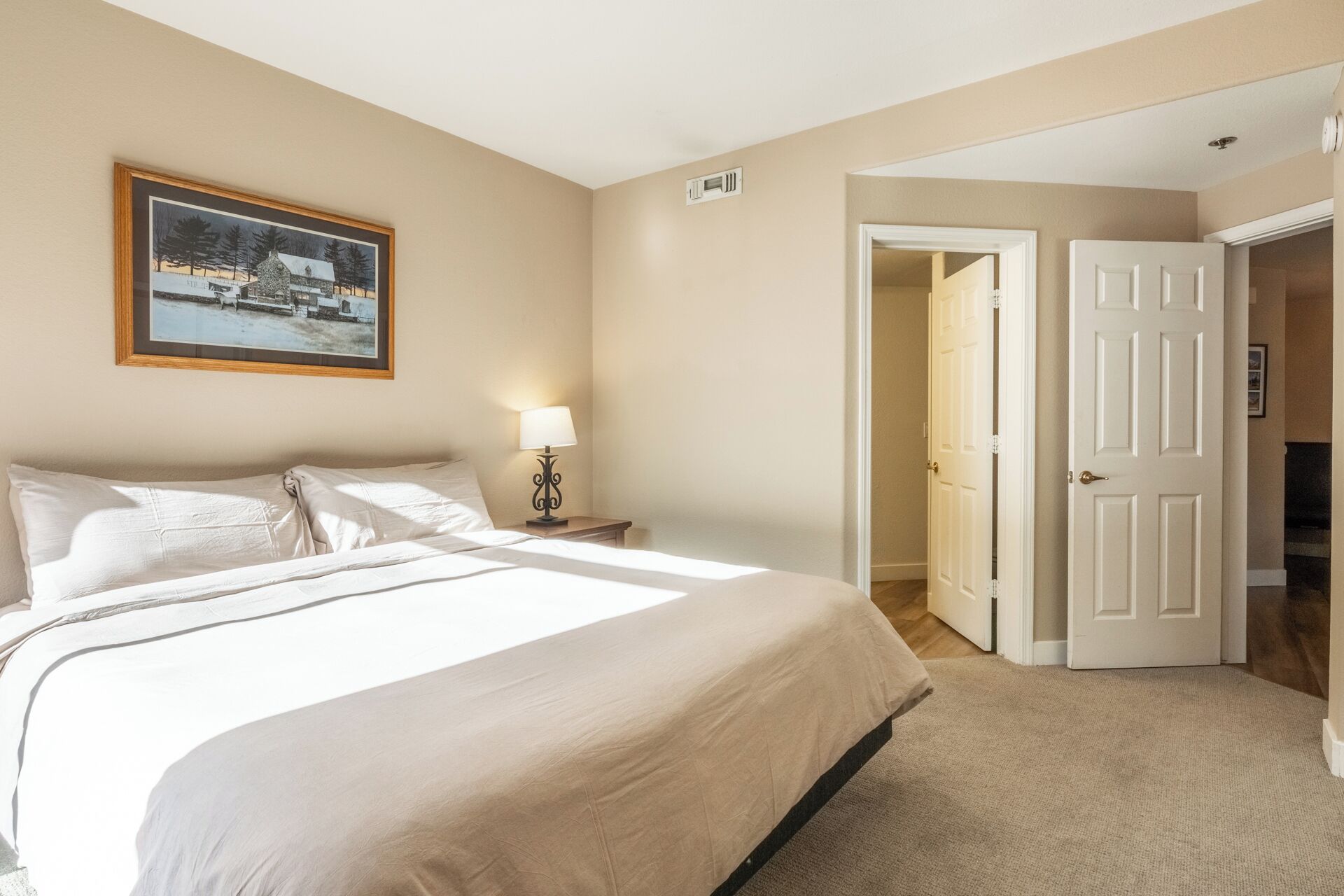 Een slaapkamer met een groot bed, een nachtkastje met een lamp, een lijst met een afbeelding aan de muur en een deur die naar een andere kamer leidt.