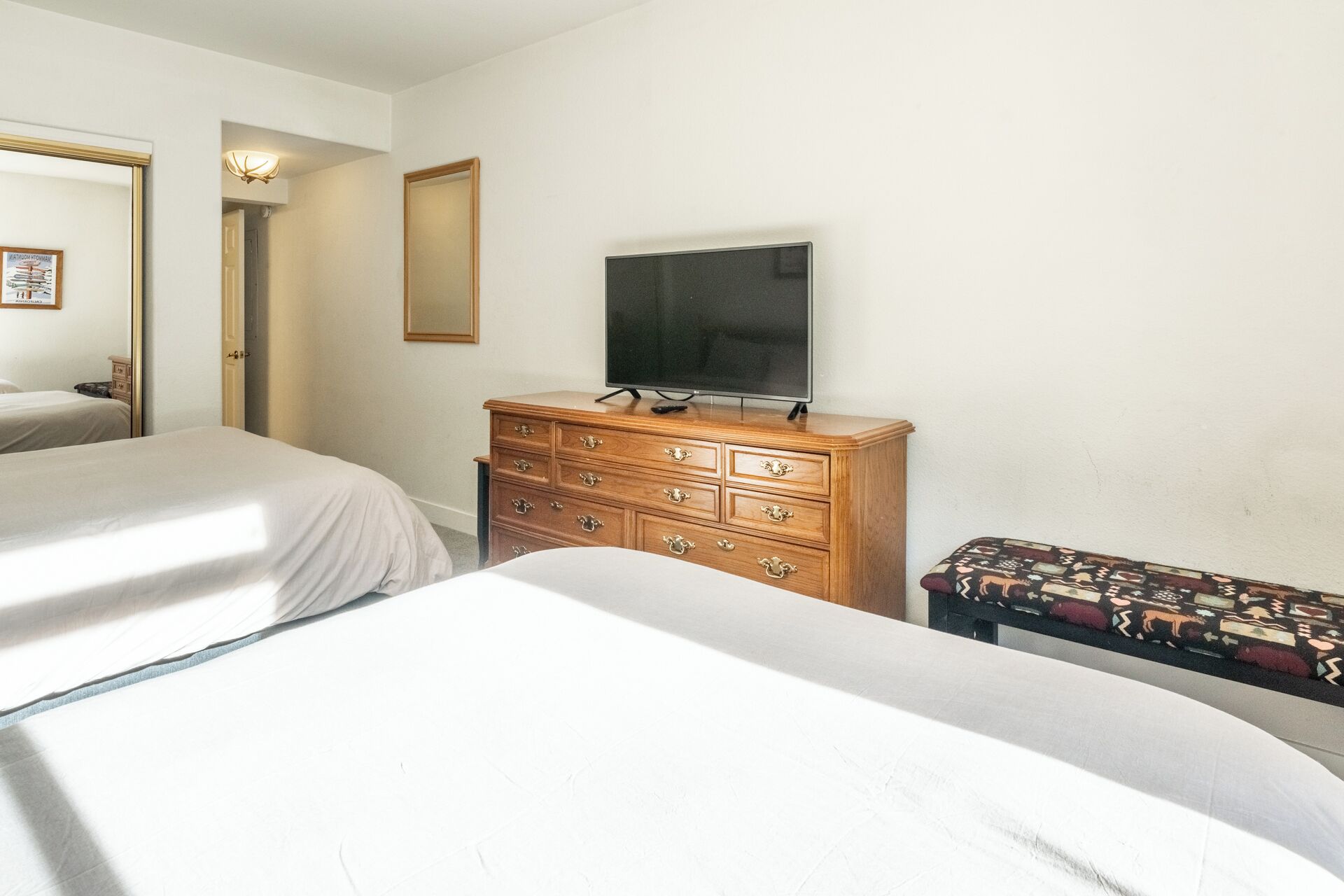 A bedroom with two beds, a wooden dresser, a television, and a patterned bench.