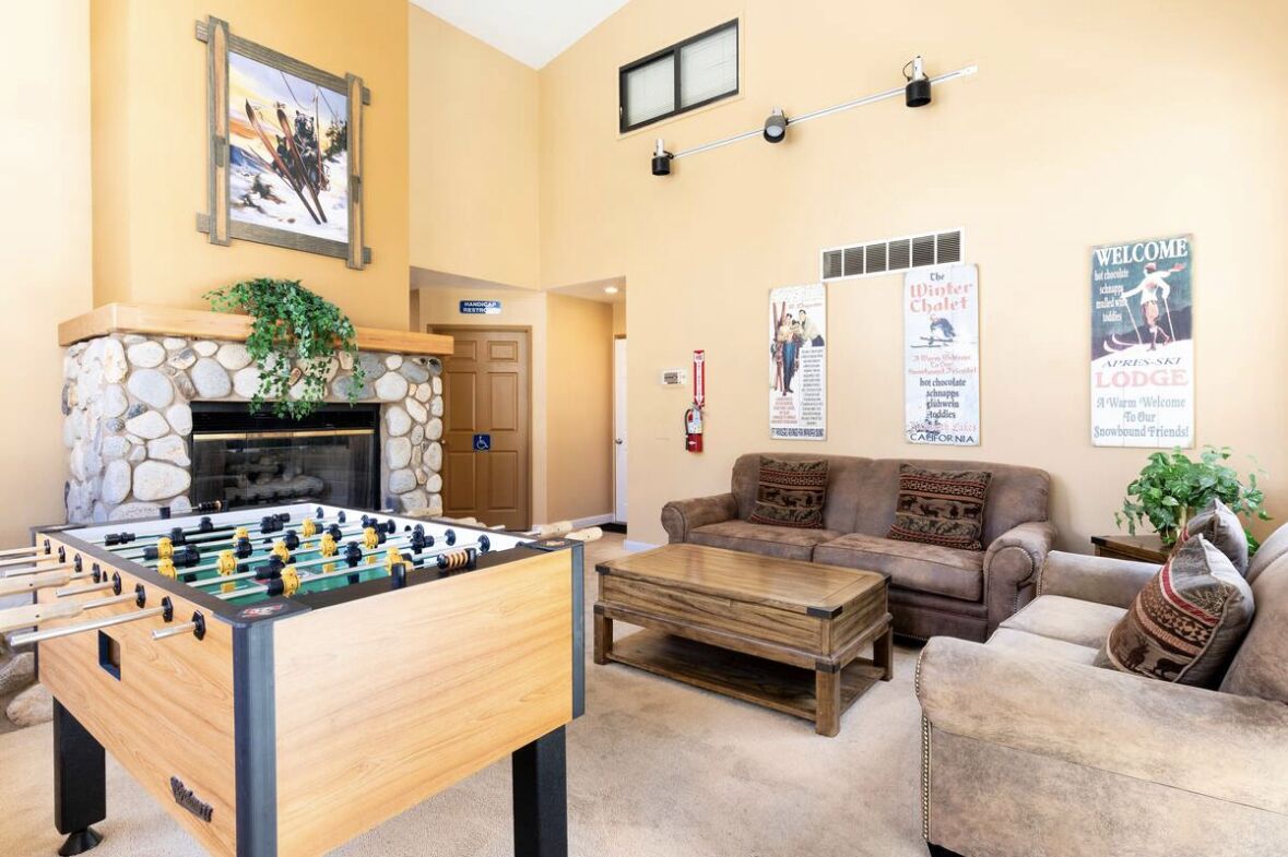 A cozy indoor area with a foosball table, a sofa, a coffee table, and a fireplace. There are posters on the wall and a sign that says "Welcome".