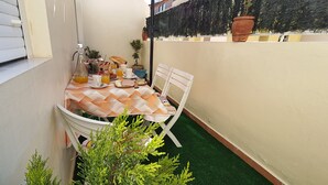 Un petit espace extérieur aménagé pour les repas, avec une table pour deux, entouré de plantes en pot et doté d’un sol recouvert d’un gazon artificiel vert.