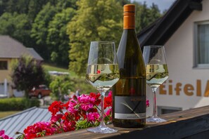 A bottle of wine and two glasses on a wooden ledge with a house and greenery in the background.