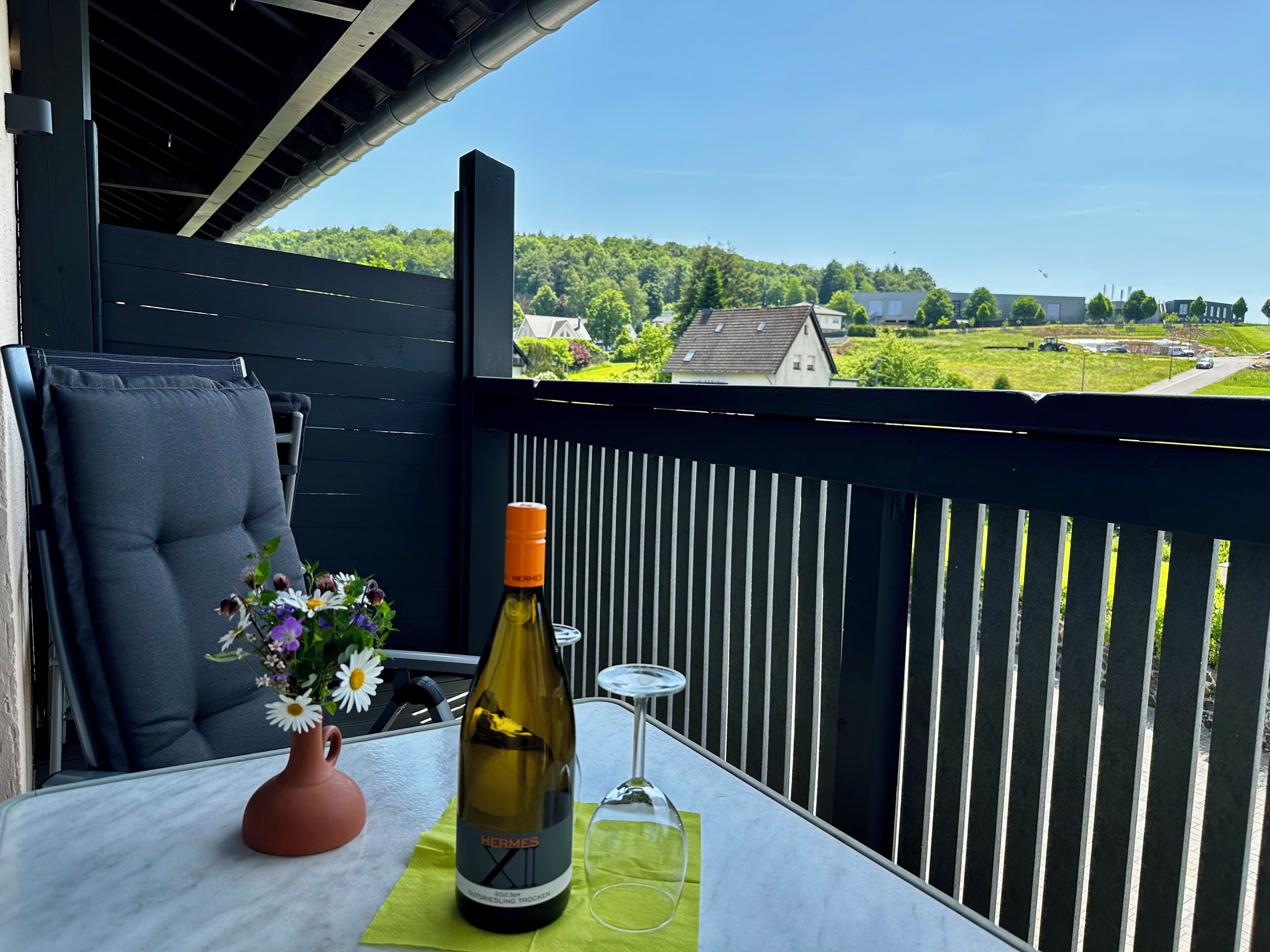 En balkong med ett bord, en flaska vin, ett glas och en vas med blommor.