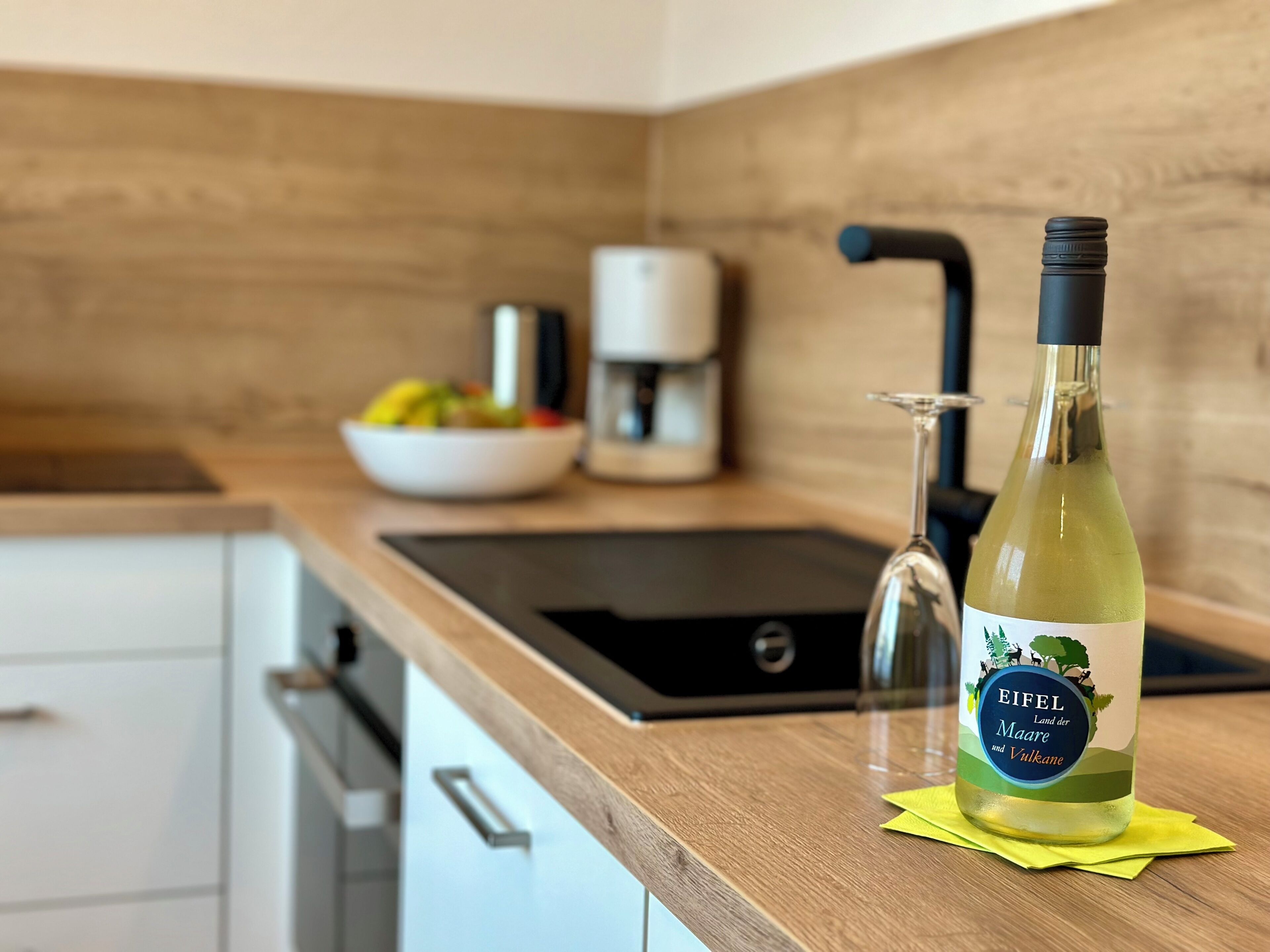 A kitchen with a sink, a coffee maker, a bowl of fruit, and a bottle of wine on the counter.