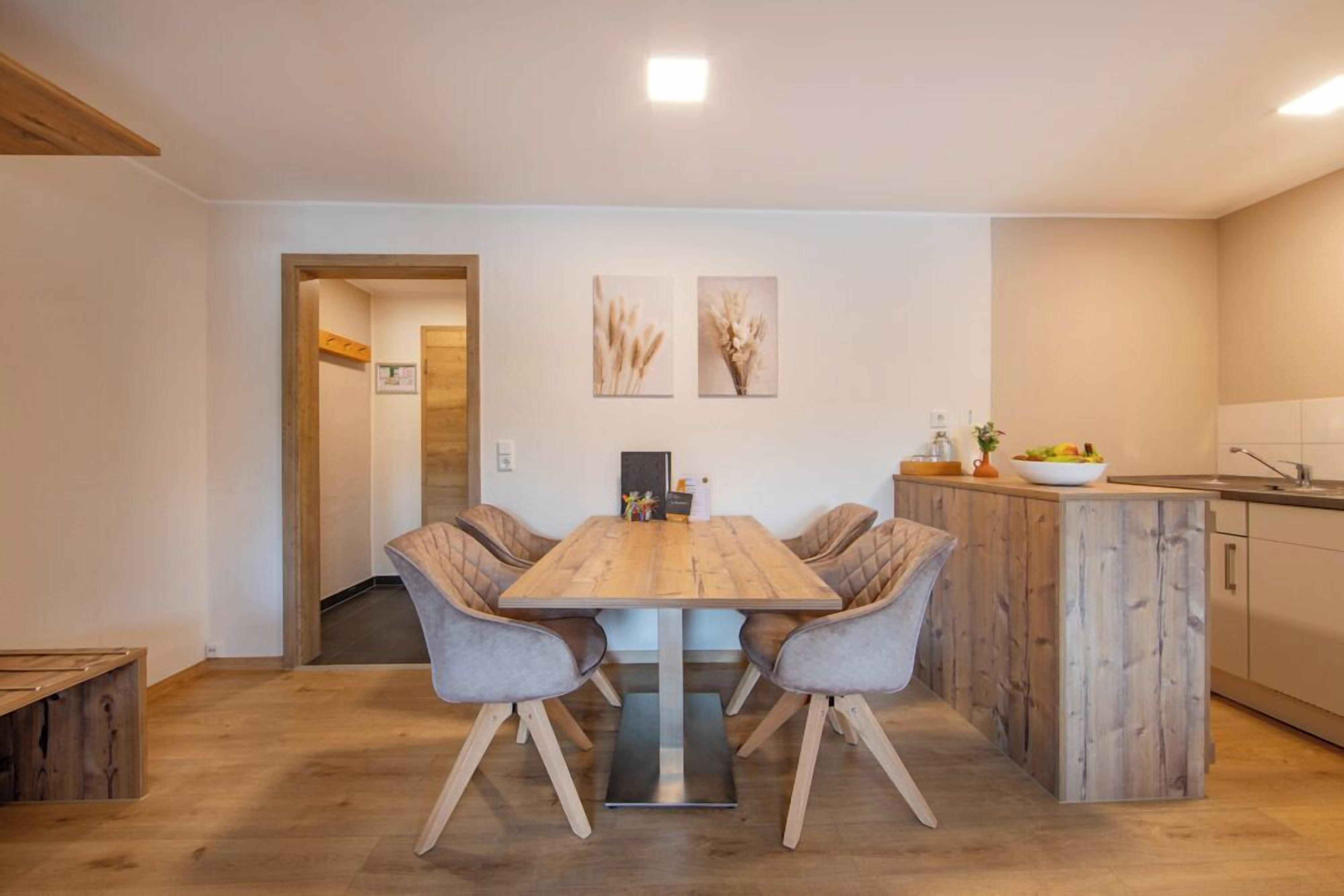 Een eetgedeelte met een houten tafel en stoelen, een keuken met een spoelbak en kasten, en twee ingelijste foto's aan de muur.