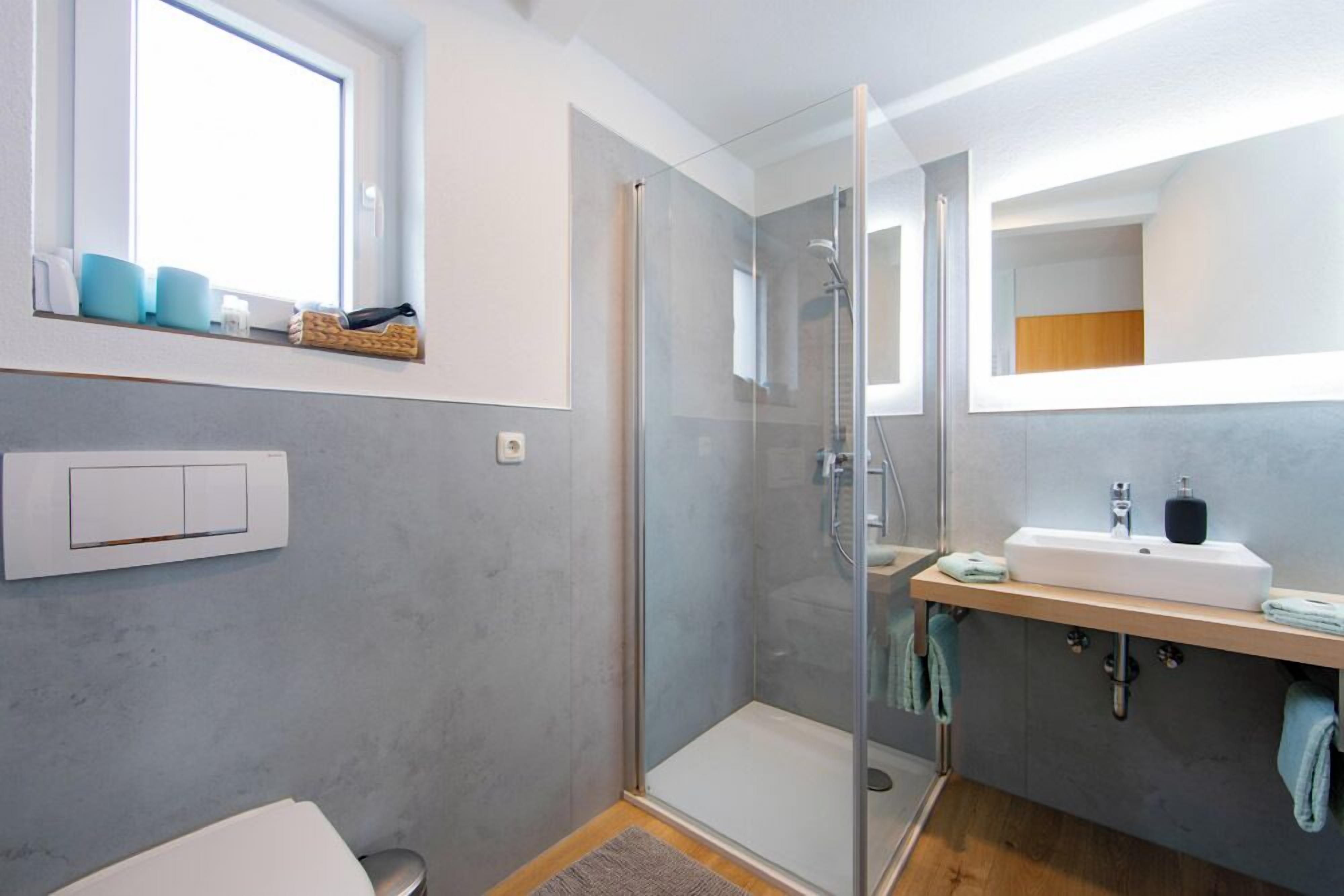A modern bathroom with a glass shower enclosure, a white sink with a wooden countertop, and a window above the sink.