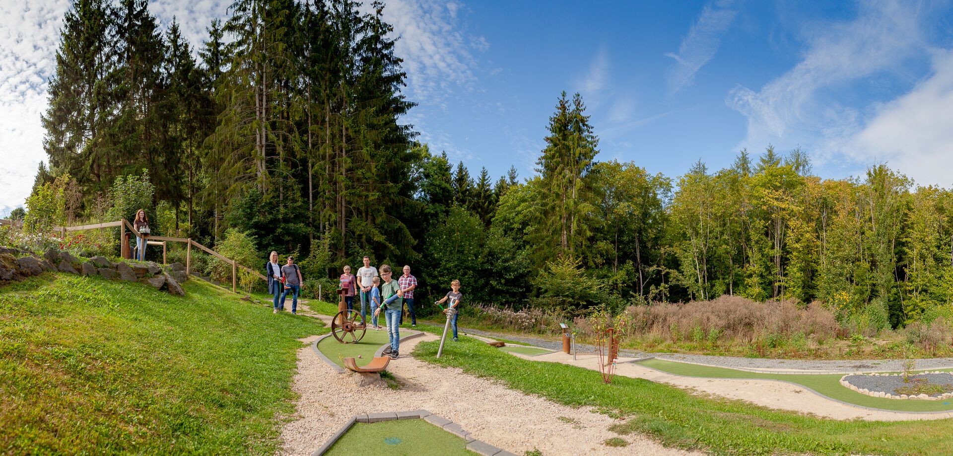A group of people playing croquet in a park with a dirt path, surrounded by trees and greenery.