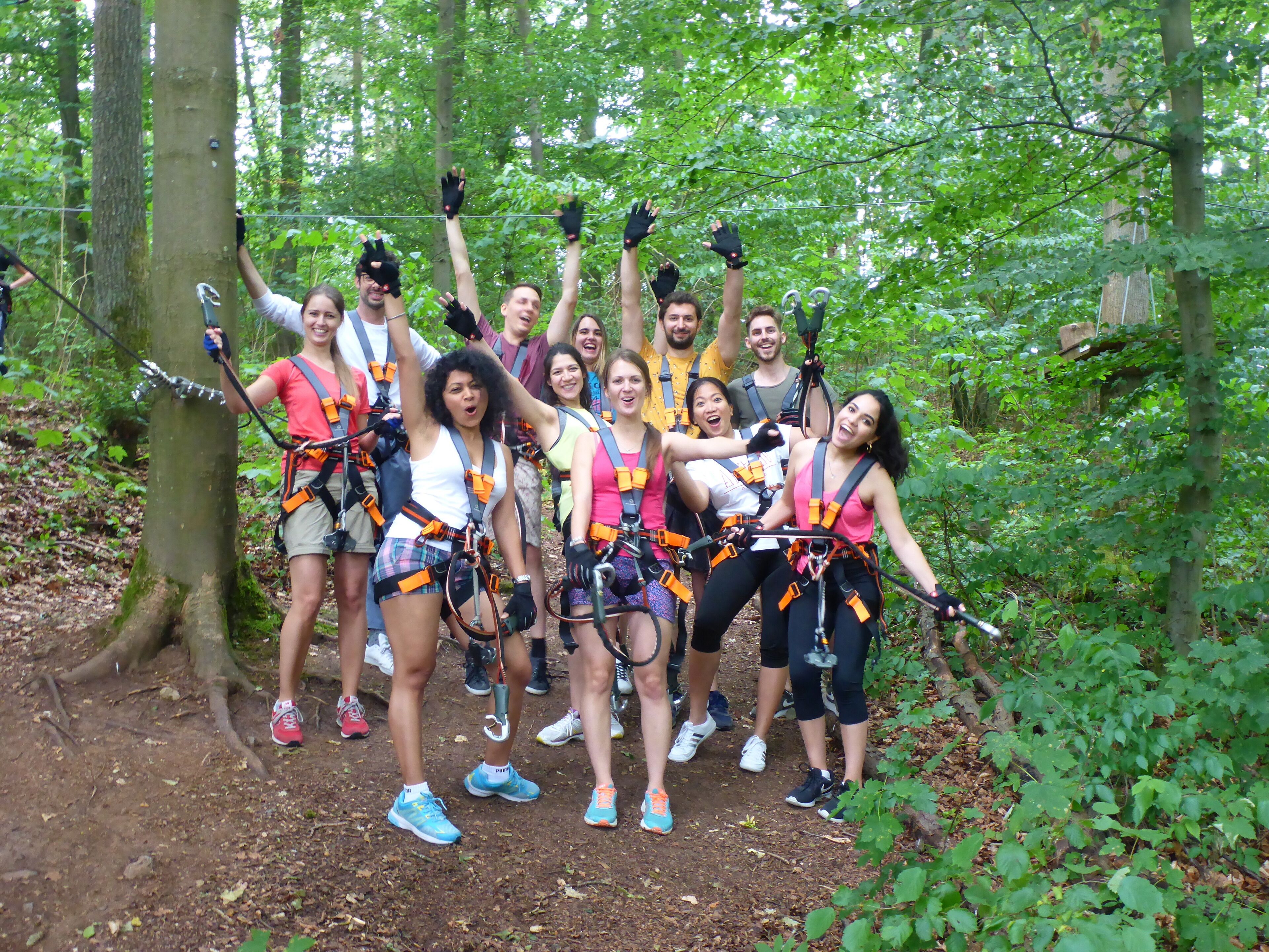 A group of people in a forested area, wearing harnesses and safety gear, likely participating in an outdoor activity.