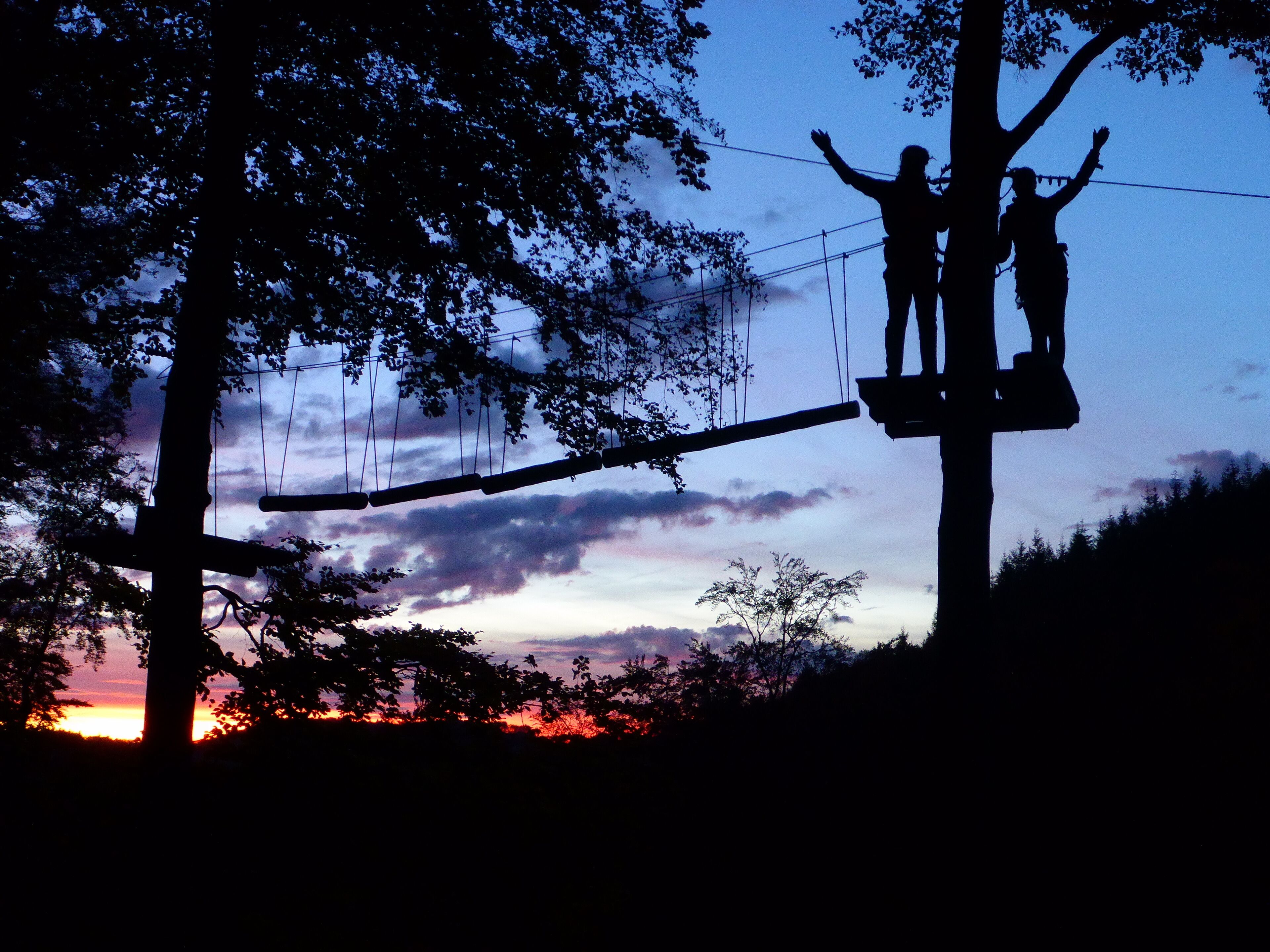 A silhouette of two people on a zip line against a sunset sky.