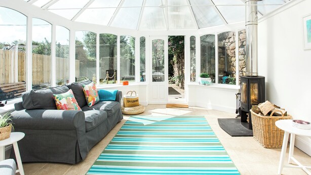 A conservatory with a grey sofa, a striped rug, a wicker basket, and a wood-burning stove. - 4 bedroom accommodation in Kirkcudbright (Kirkcudbright)