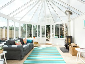 A conservatory with a grey sofa, a striped rug, a wicker basket, and a wood-burning stove. - 4 bedroom accommodation in Kirkcudbright (Kirkcudbright)
