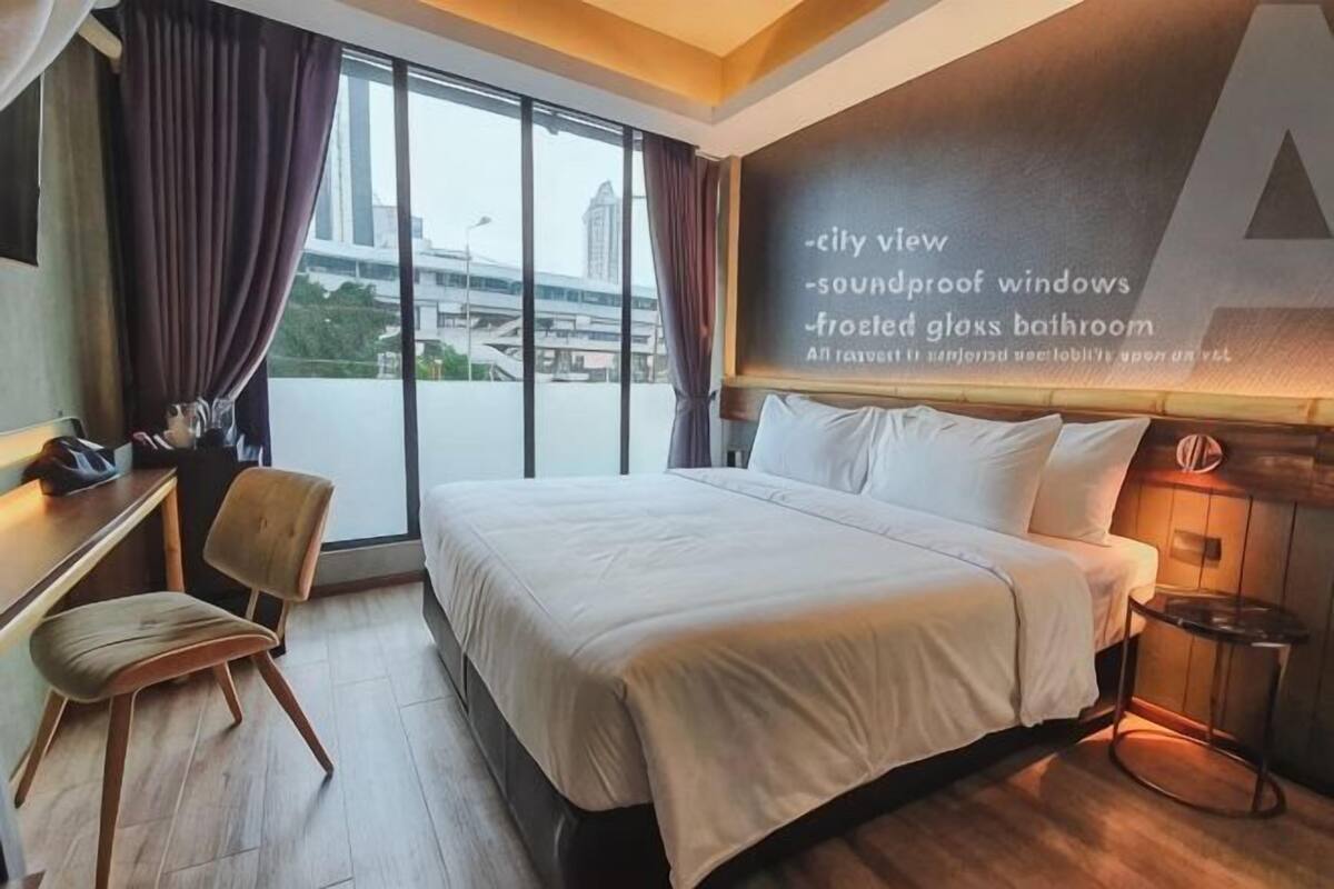 A hotel room with a bed, a desk, a chair, a window with curtains, and a wall-mounted screen displaying amenities.
