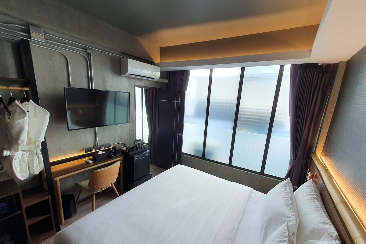 A hotel room with a large bed, a desk with a computer, a TV, and a view of the city through large windows.