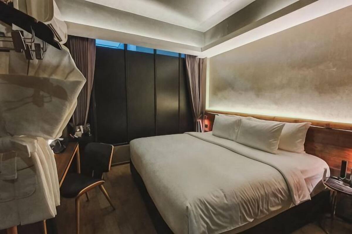 A hotel room with a large bed, a desk with a chair, a wardrobe, and a nightstand with a lamp.