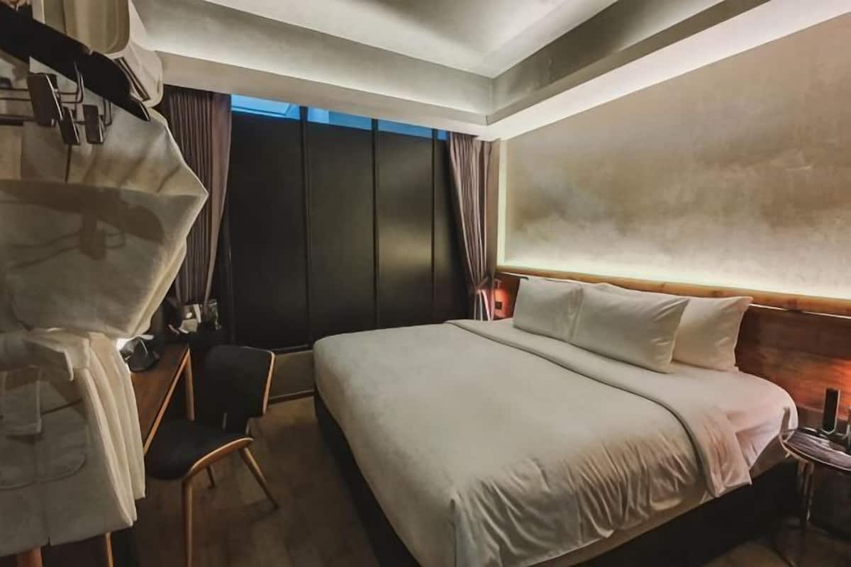 A hotel room with a large bed, a desk with a chair, a wardrobe, and a nightstand with a lamp.