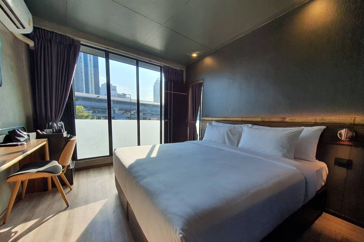 A hotel room with a large bed, a desk, and a chair. There is a view of the city through the windows.