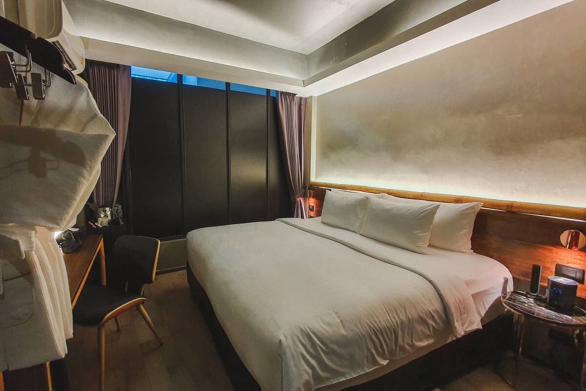 A hotel room with a large bed, a desk with a chair, a small table with a lamp, and a wardrobe.