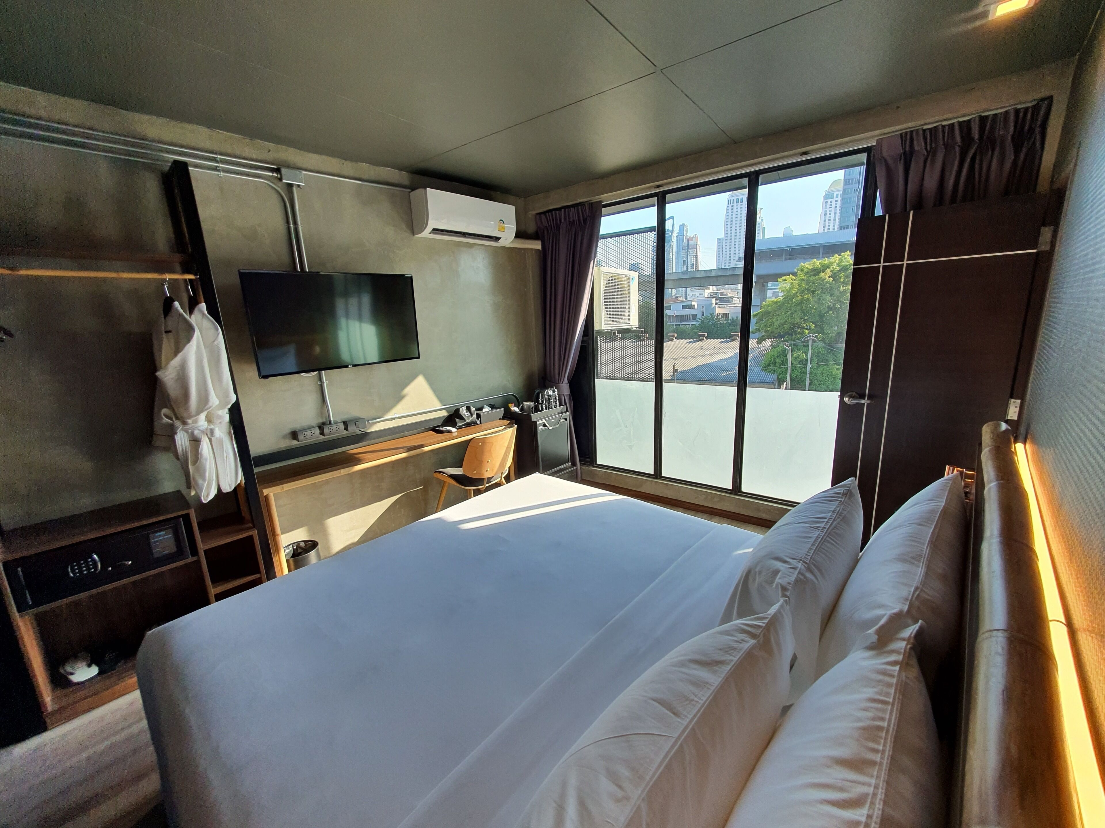 A hotel room with a large bed, a desk with a chair, a TV mounted on the wall, and a window with a city view.