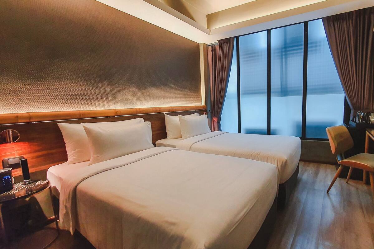 A hotel room with two beds, a desk, a chair, and a large window with curtains.