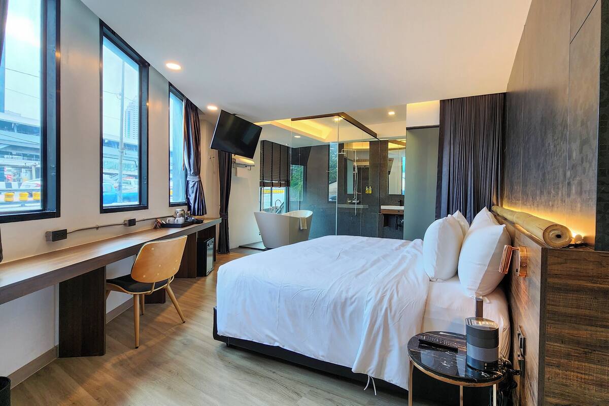 A modern hotel room with a large bed, a desk, a chair, a flat-screen TV, and a bathroom with a mirror and sink.