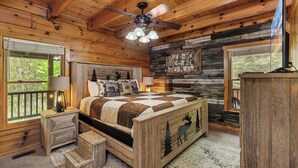 A wooden cabin bedroom with a bed, a nightstand, a lamp, a ceiling fan, and a window with a view of trees. - Free Tickets | Creek-Side, Views, INSANE Arcade, Yard, Fireplace (Sevierville)