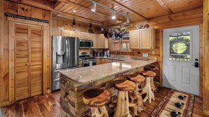 A cabin kitchen with wooden walls, a granite island with stools, stainless steel appliances, and a welcome sign. - Free Tickets | Creek-Side, Views, INSANE Arcade, Yard, Fireplace (Sevierville)