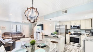 Una cocina moderna con armarios blancos, electrodomésticos de acero inoxidable y un comedor con mesa y sillas de vidrio.