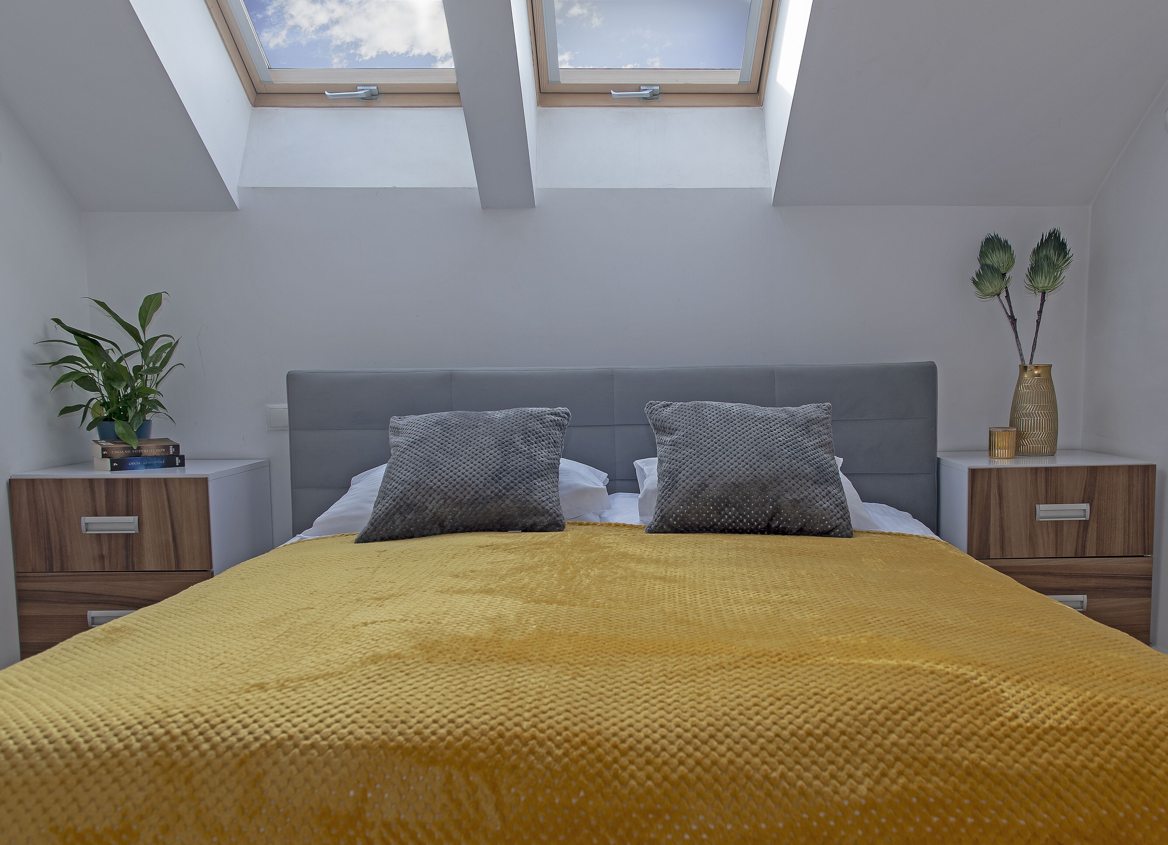 Een slaapkamer met dakraam, een bed met een geel geweven deken, twee kussens, een kamerplantje en een nachtkastje met een lade.