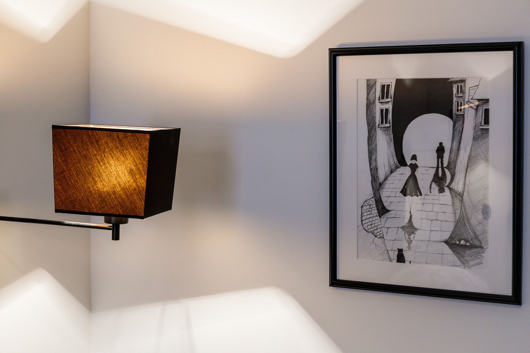 Une reproduction encadrée en noir et blanc représentant une rue étroite où l’on voit une femme et un homme, une lampe murale moderne et un mur blanc.