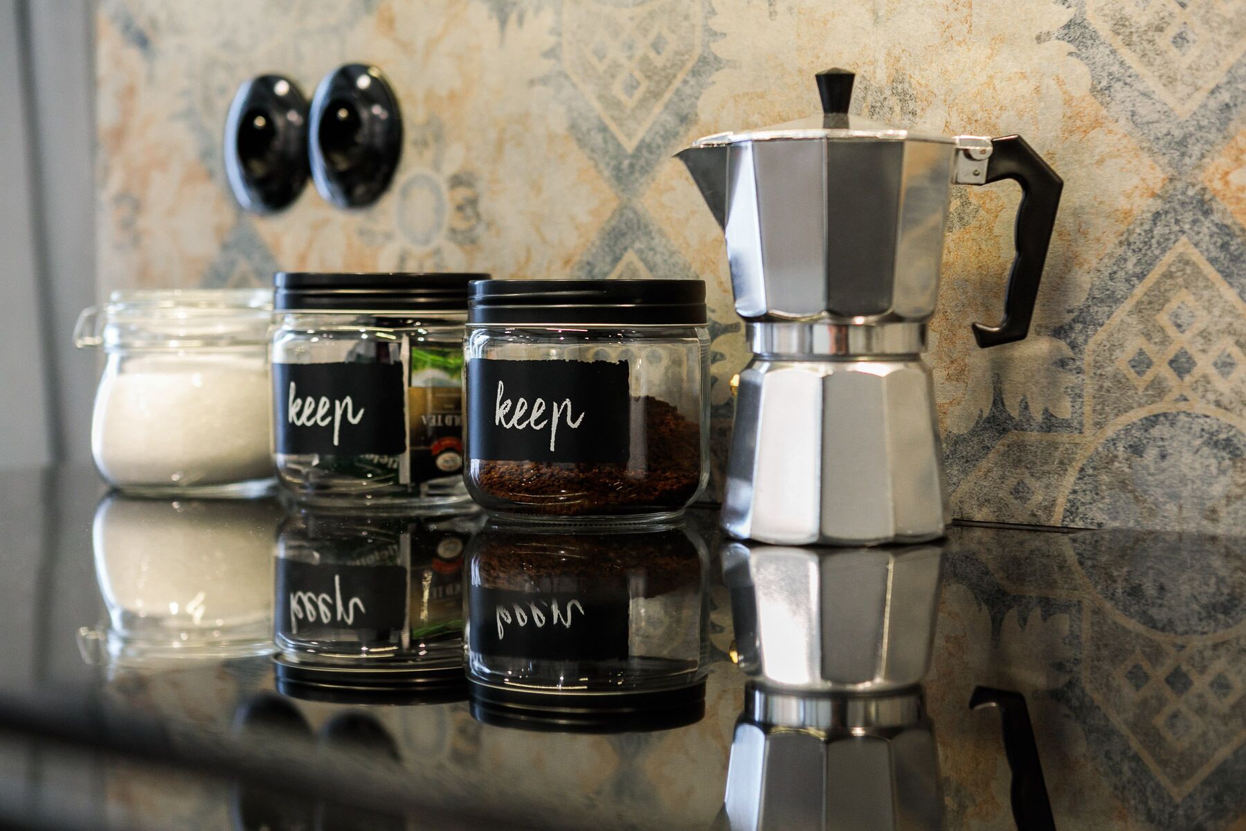 Three glass jars with black lids labeled "keep" on a reflective countertop, next to a silver coffee pot.