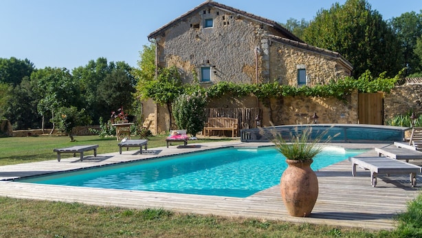 Un edificio de piedra con piscina, terraza de madera y sillones de descanso.