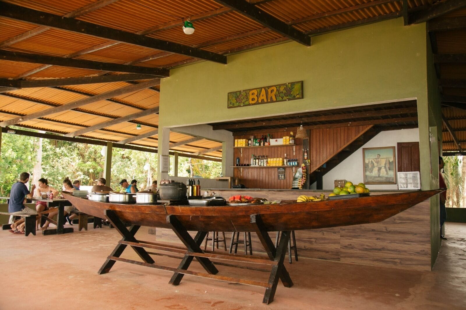 A rustic bar area with wooden beams and a green wall, featuring a long wooden table and a bar counter with various bottles and a fruit display.