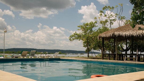 A swimming pool with lounge chairs and a thatched-roof structure, surrounded by trees and a view of a body of water. - Anaconda Amazon Resort (Manaus)