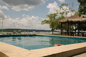 A swimming pool with lounge chairs and a thatched-roof structure, surrounded by trees and a view of a body of water. - Anaconda Amazon Resort (Manaus)