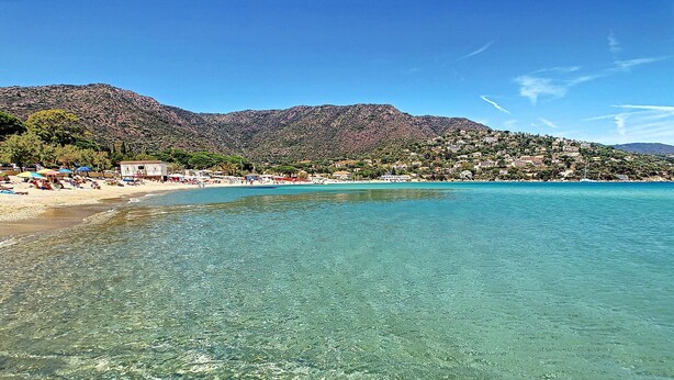 A clear beach with turquoise water, a sandy shore, and a mountainous backdrop.