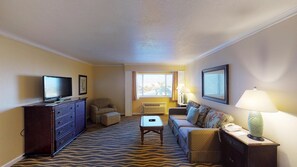 A hotel room with a television, a sofa, a patterned carpet, a window with curtains, and a lamp.