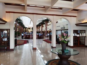 Um hall amplo com pé direito alto, uma mesa de vidro central e uma área de recepção.