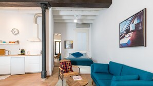 Ein modernes Wohnzimmer mit einem Türkis-farbenen Sofa, einem Holztisch und einem Korbstuhl. Im Hintergrund ist eine Küche mit weißen Schränken und einer Uhr erkennbar.