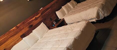 Quarto com duas camas de solteiro, cabeceira de madeira, ar condicionado fixado na parede e uma mesa pequena com luminária.