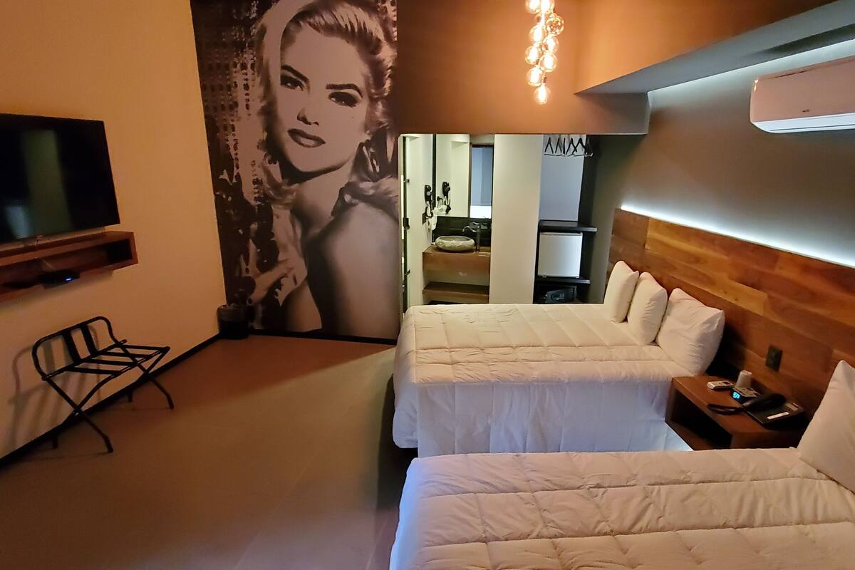 A hotel room with two beds, a TV, a wall-mounted air conditioner, and a seating area with a chair.