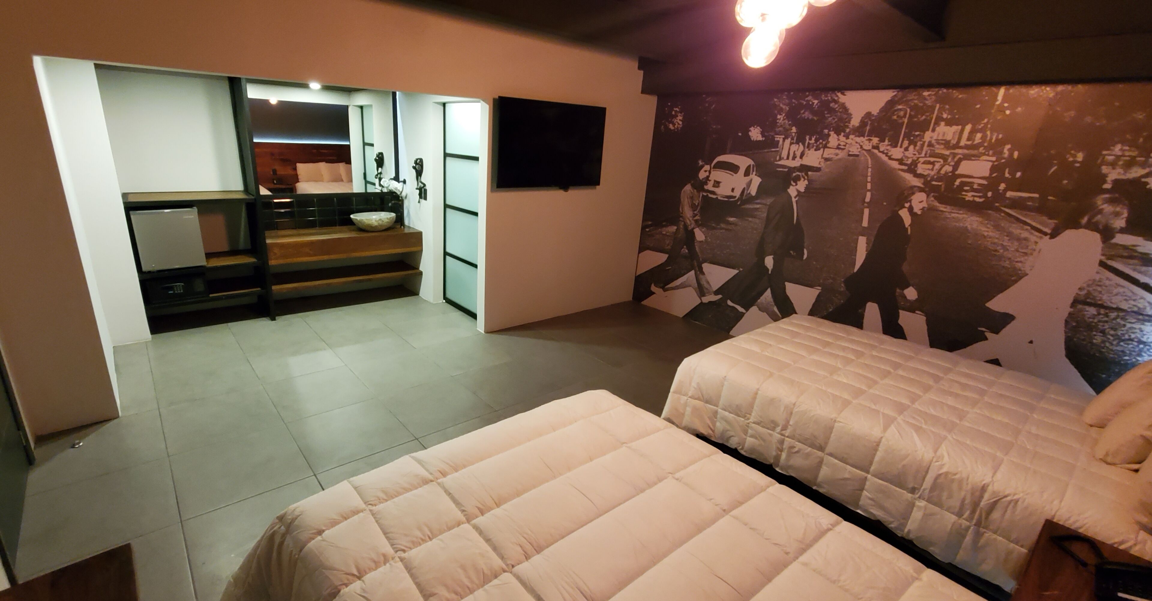Habitación de hotel con dos camas, una kitchenette pequeña, un baño con espejo y un televisor de pared.