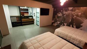 Habitación de hotel con dos camas, una kitchenette pequeña, un baño con espejo y un televisor de pared.