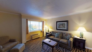 A living room with a sofa, a coffee table, and a side table with lamps. There is a window with curtains, a radiator, and a framed picture on the wall.