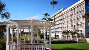 A white gazebo with a red floor, surrounded by a white fence, in front of a multi-story hotel with balconies and palm trees. - St Pete Beach | Couples Gulf Coast Retreat | Pool + Beach (St. Pete Beach)