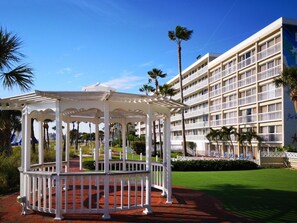 A white gazebo with a red floor, surrounded by a white fence, in front of a multi-story hotel with balconies and palm trees. - St Pete Beach | Couples Gulf Coast Retreat | Pool + Beach (St. Pete Beach)
