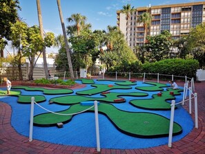 Un campo de mini golf con superficie verde y azul, bordeado por ladrillos y palmeras.